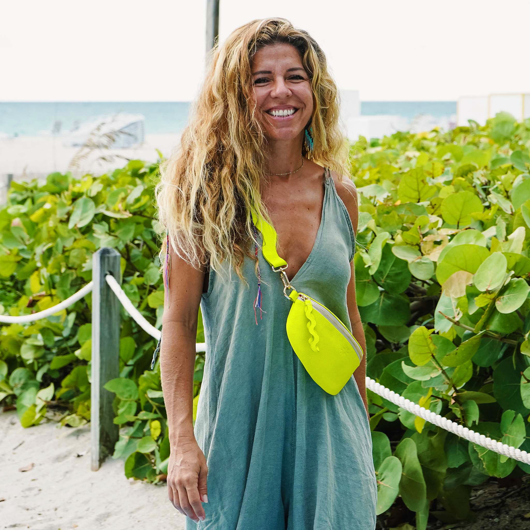 Woman in a teal dress with a neon green bag standing on a beach path.