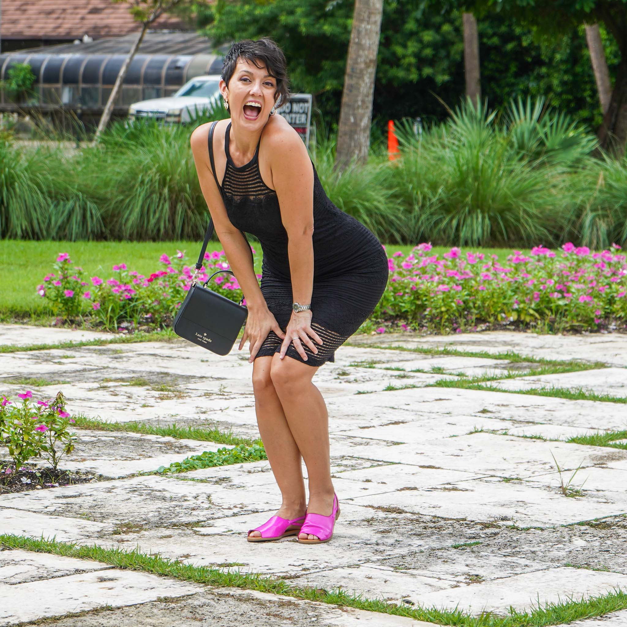 Woman in a black dress with pink shoes holding a black handbag on a stone path with greenery and flowers in the background.