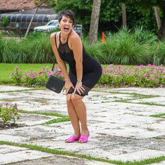 Woman in a black dress with pink shoes holding a black handbag on a stone path with greenery and flowers in the background.